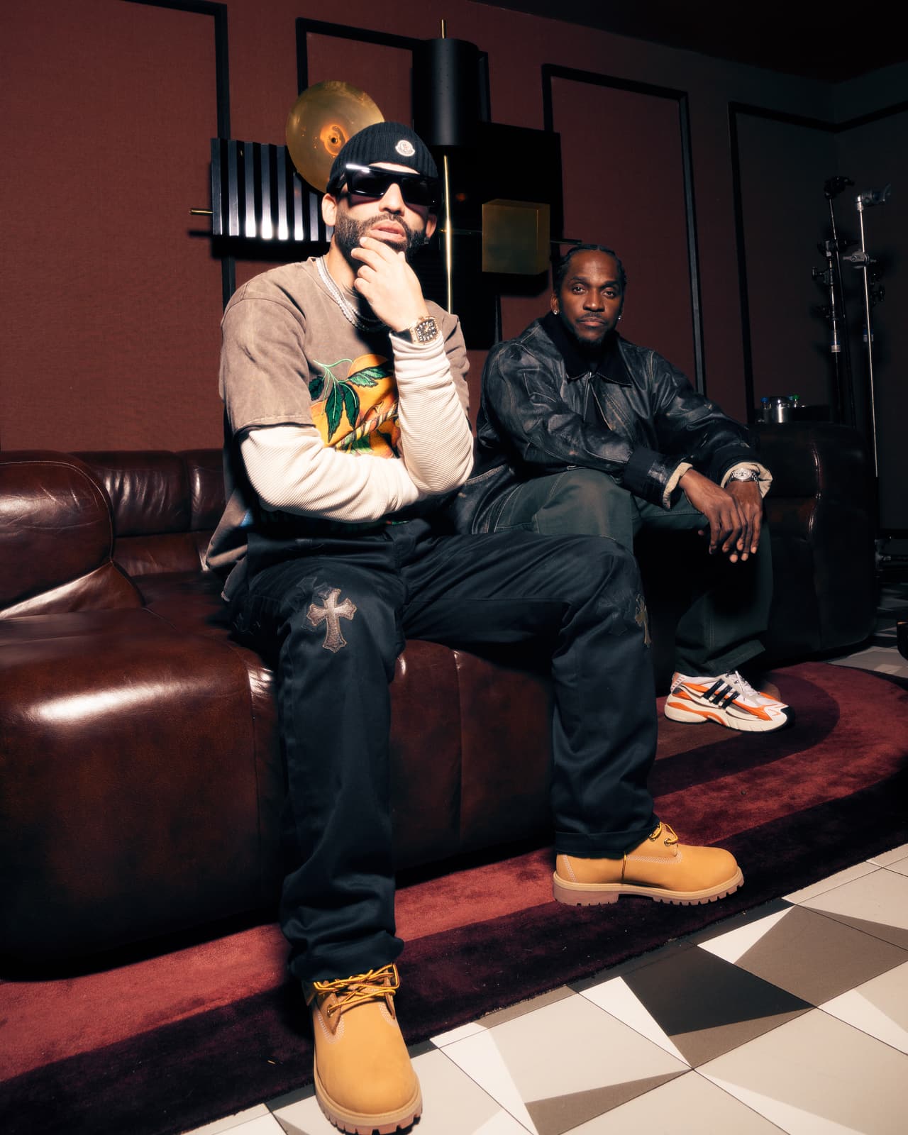Two men in an oxblood lounge, one perched on the sofa arm in sunglasses and a printed crewneck, one seated behind in a distressed leather jacket