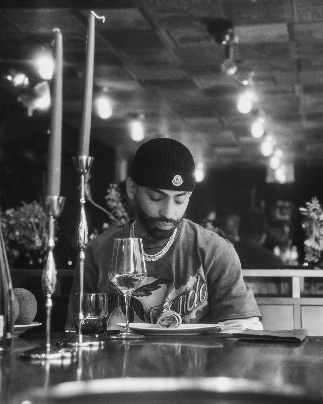 Black-and-white portrait at a dining table, a printed crewneck and a tennis chain, a run of bare bulbs receding behind