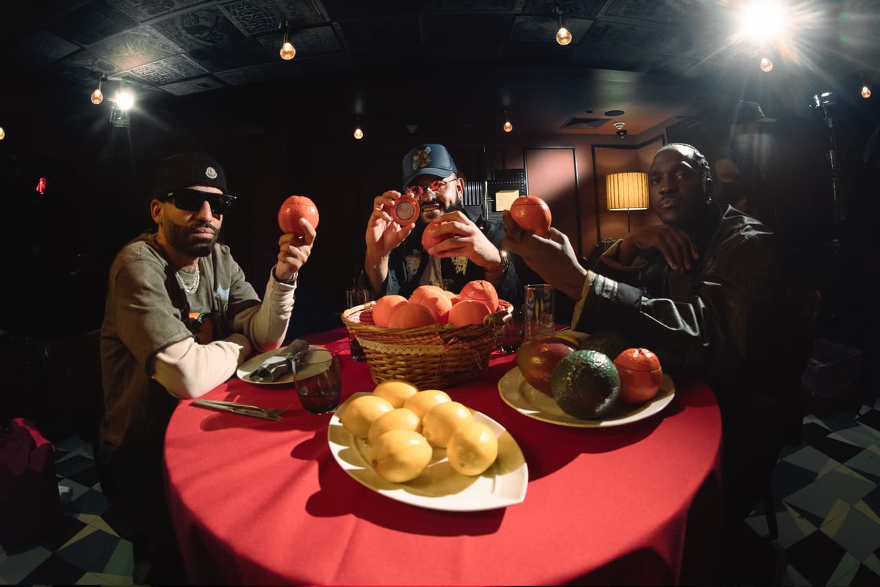 Three men across a red table each holding a blood orange up to the lens, one of them also holding a lid with an embossed disc