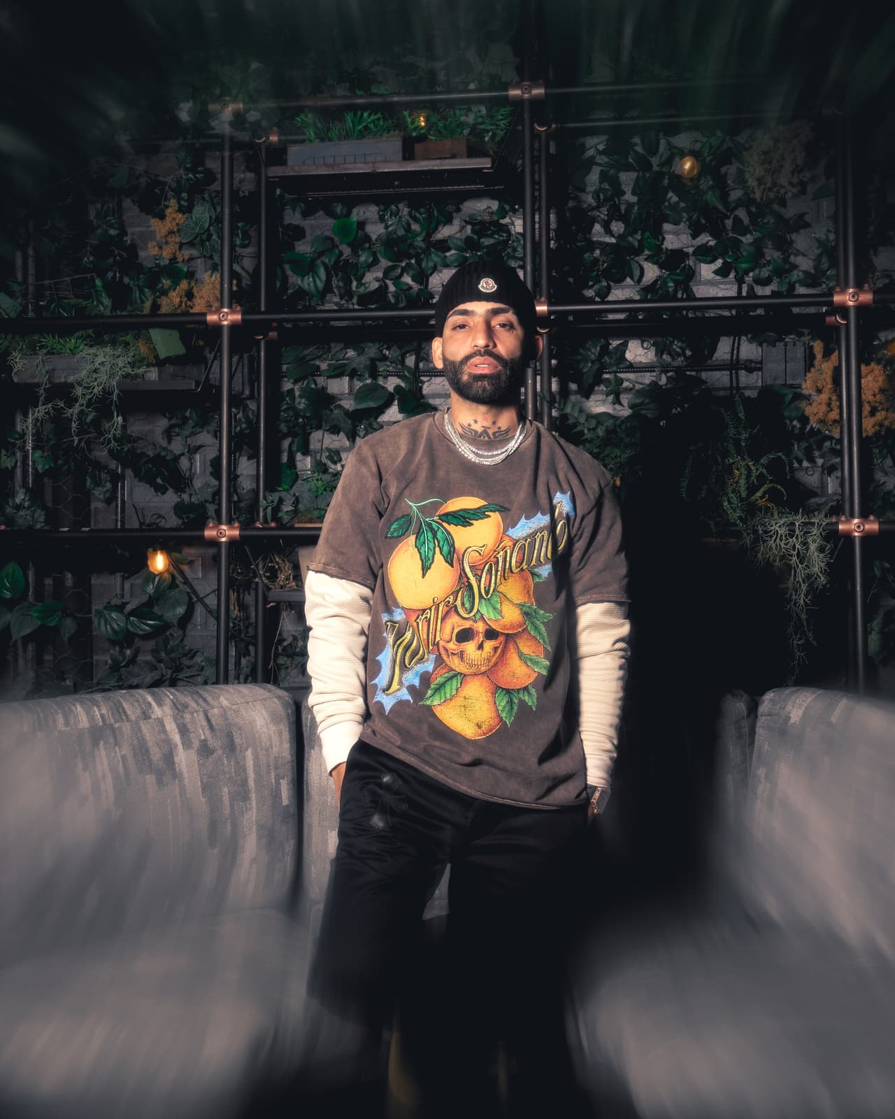 A man standing square to camera on a planted terrace, hands in pockets, the full oranges-and-skull crewneck print unobstructed