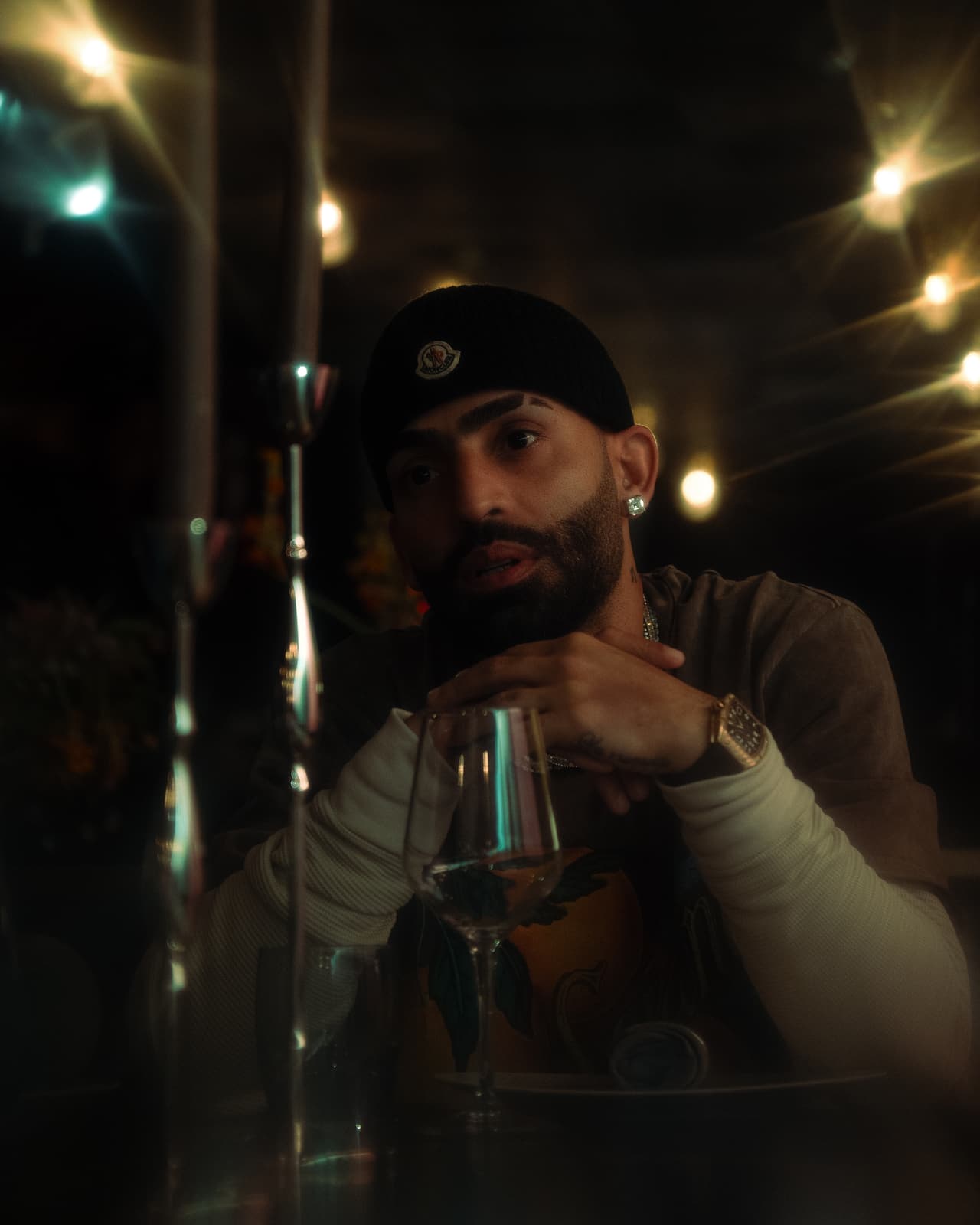 A man at a laid dining table with his hands crossed over a wine glass, bare-bulb string lights starring behind him