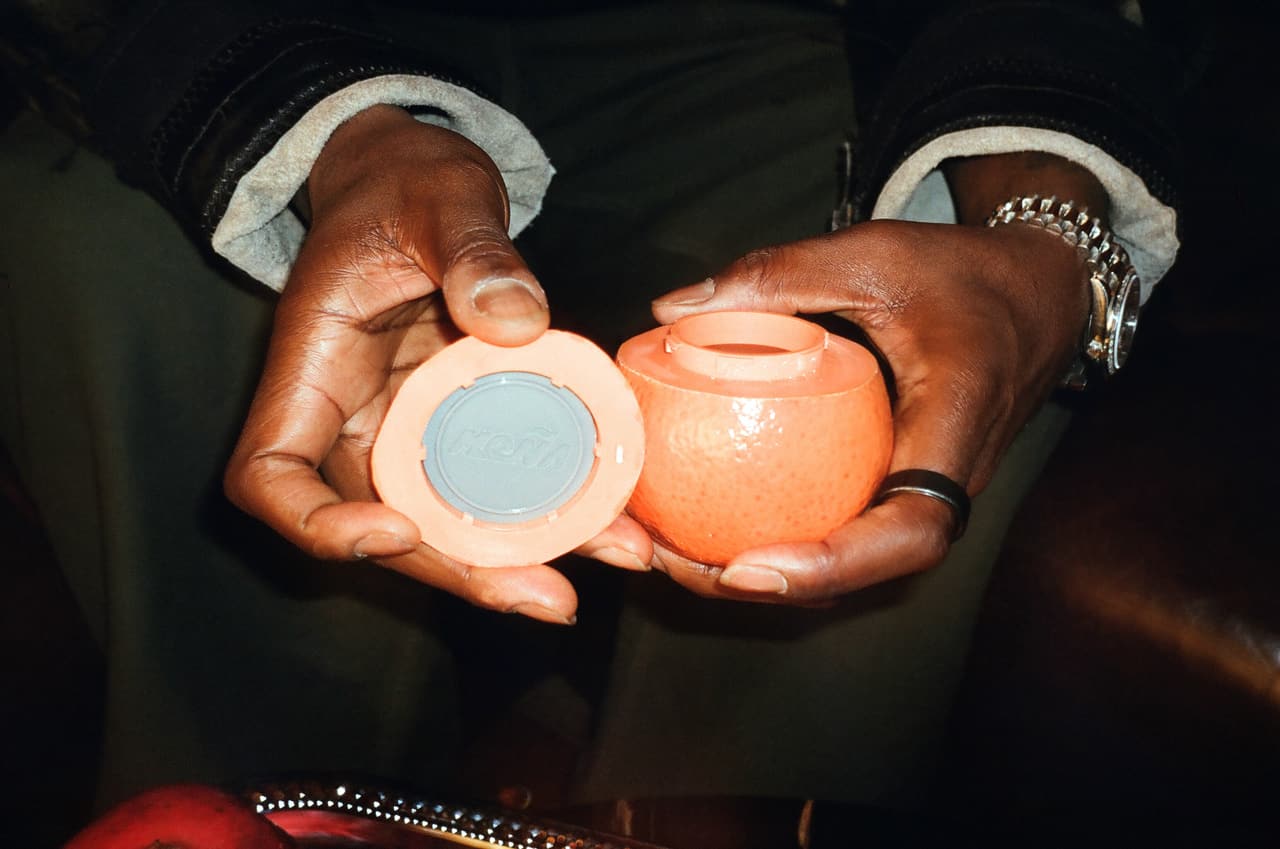 Two hands presenting an opened ceramic orange, the lid held flat in one palm with a MOÑA-embossed disc set into it