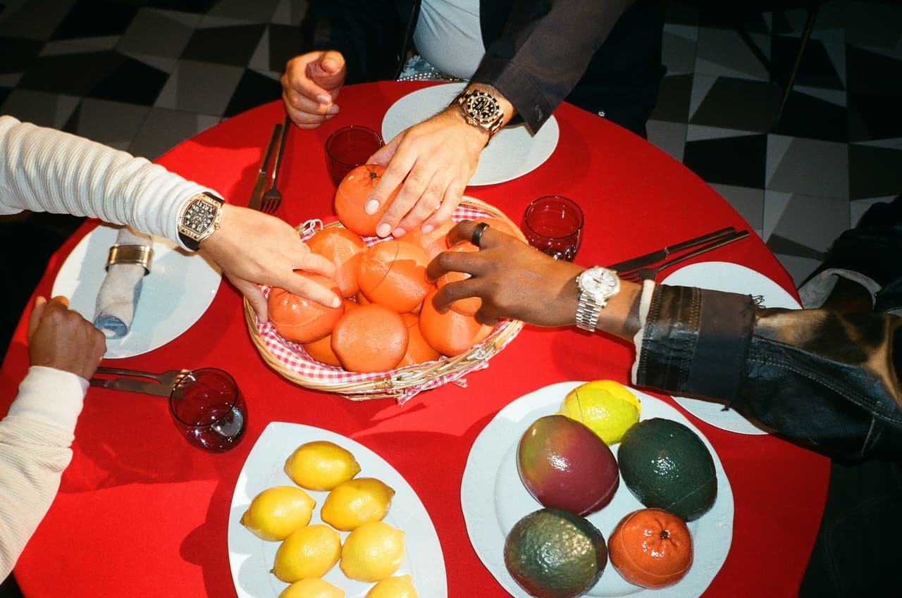 Overhead of four hands reaching into a gingham-lined basket of oranges on a red cloth, ruby glasses and gold cutlery around it