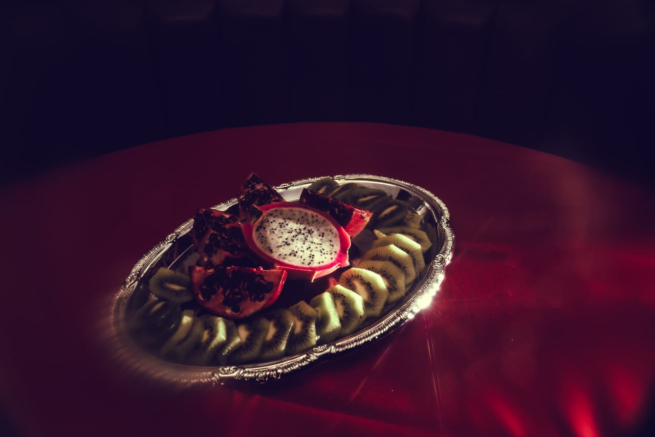 A silver oval platter of halved dragon fruit, quartered pomegranate and fanned kiwi, spot-lit on a red tablecloth against near-black