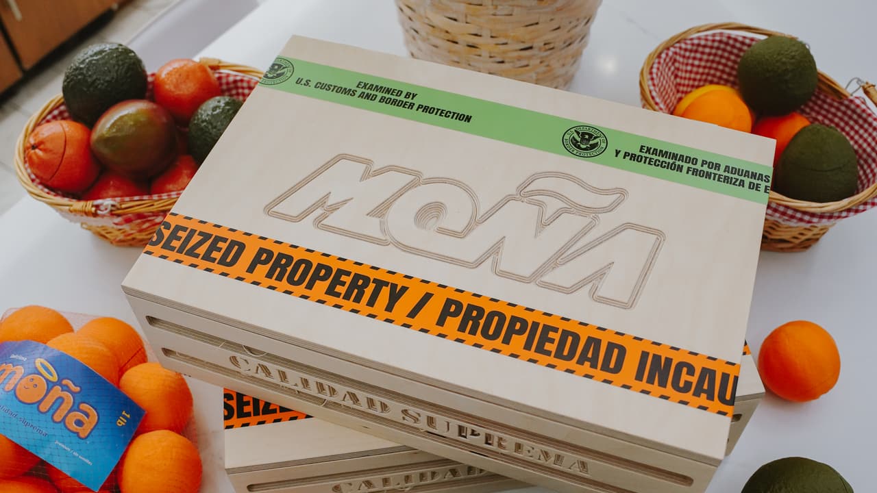 Top-down view of a MOÑA seized-property crate with orange and avocado net bags and fruit baskets on white marble