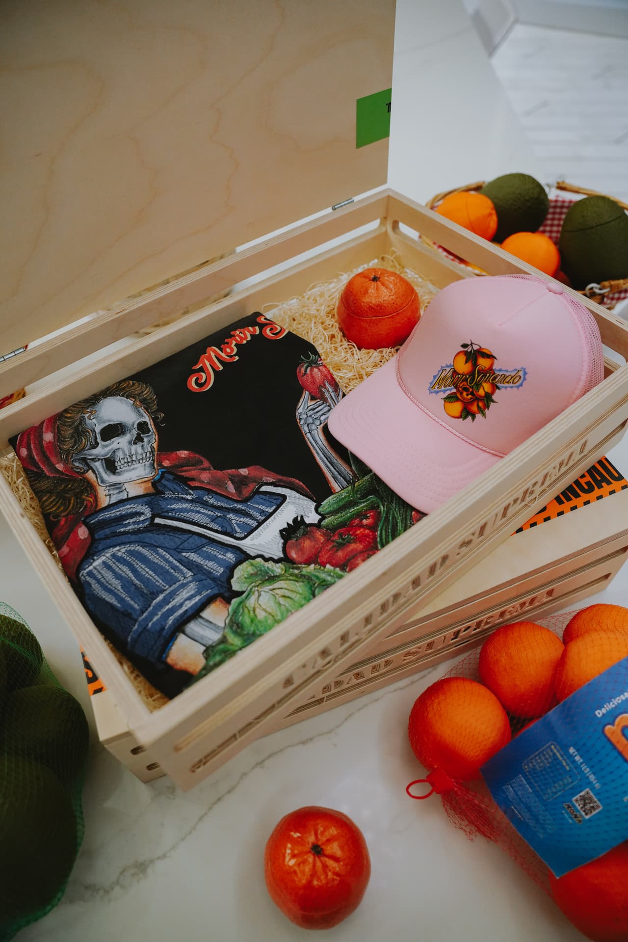 Open crate with a folded black harvest tee, a pink trucker hat and a ceramic orange prop on wood wool packing