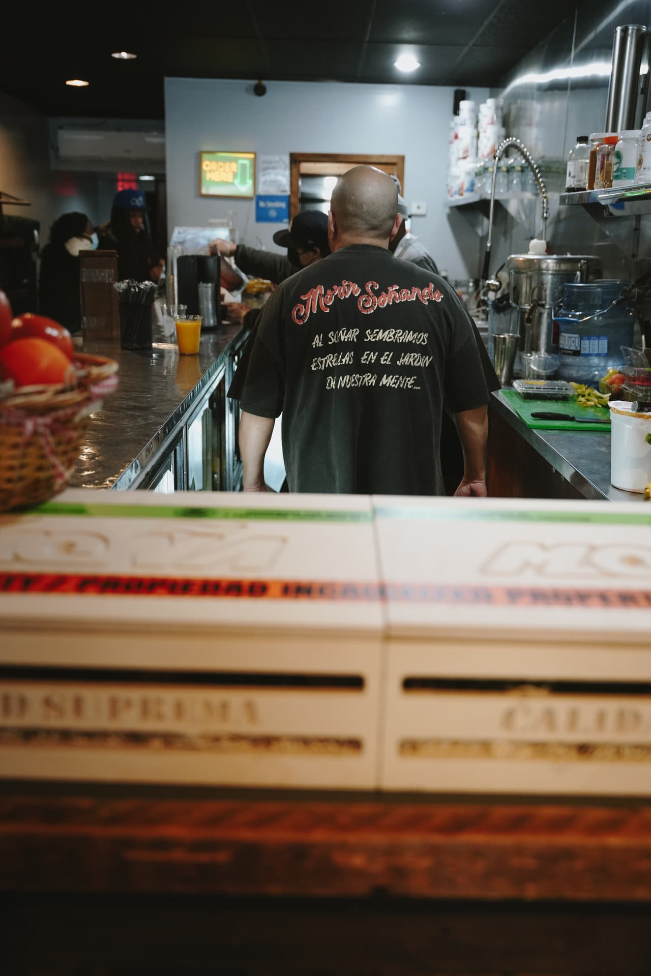 Back of the harvest tee reading 'Morir Soñando / Al soñar sembramos estrellas en el jardín de nuestra mente', crates and baskets in the foreground