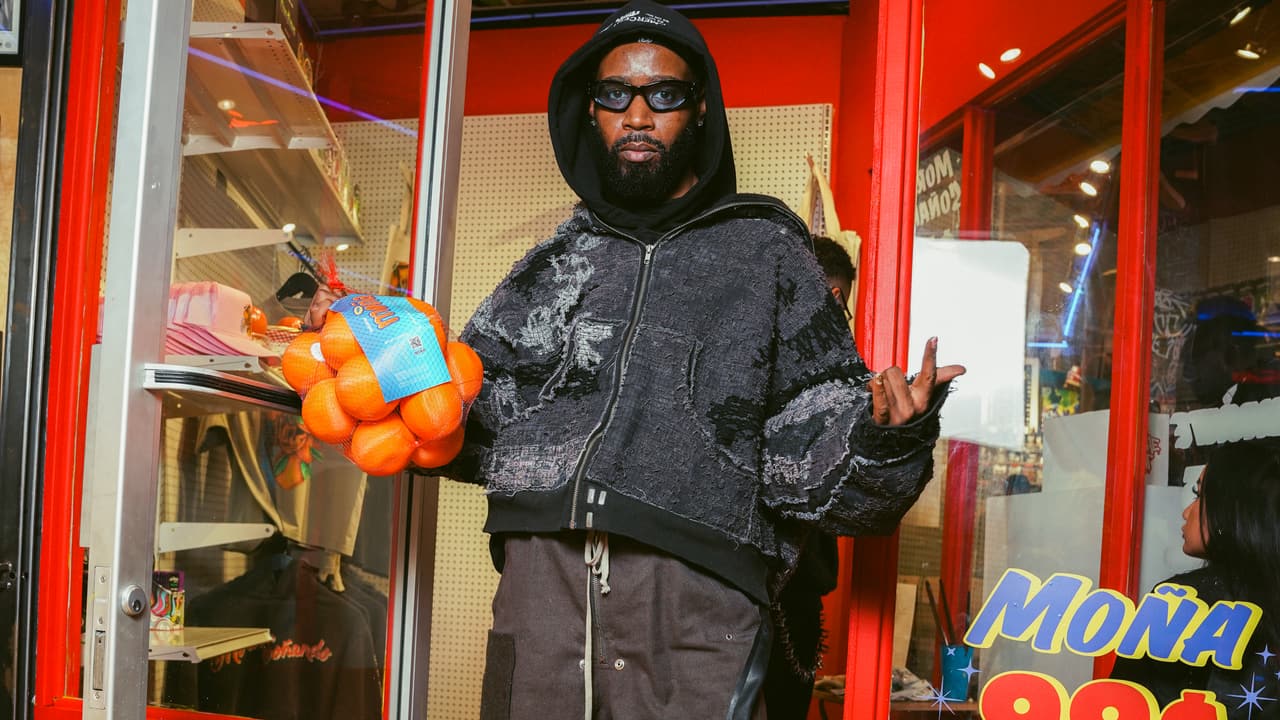 A guest in a black hood and sunglasses holding a MOÑA orange net bag, standing in a symmetrical kiosk doorway under a 'MOÑA 99¢' sign
