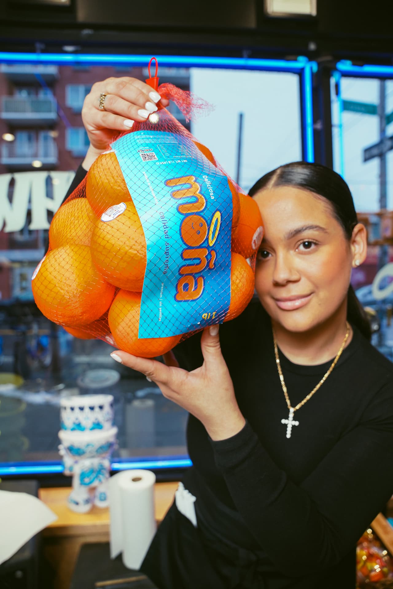 A guest holding a MOÑA orange net bag to her cheek, a blue neon window frame behind her