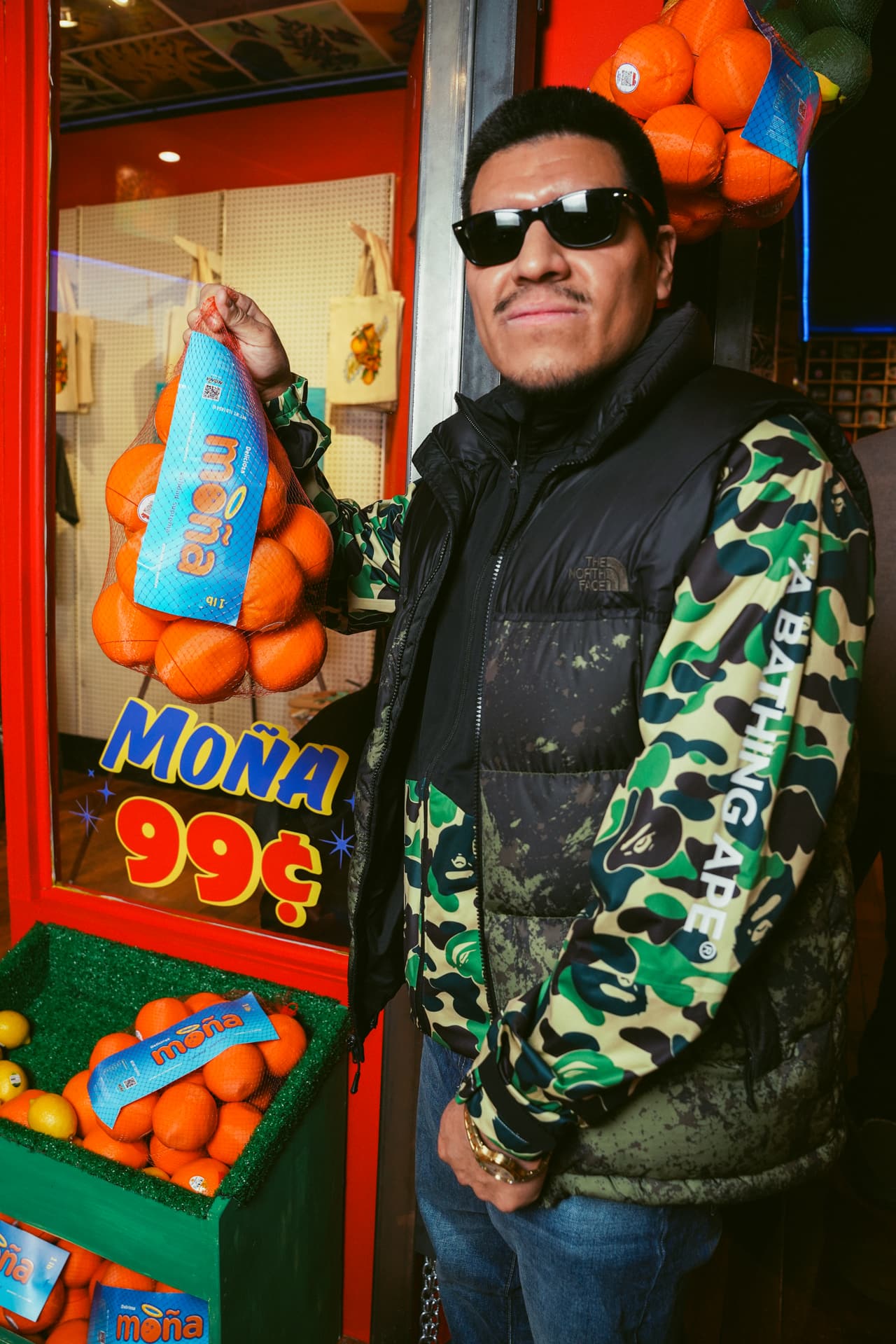 A guest holding a MOÑA orange net bag at the 'MOÑA 99¢' window, a crate of oranges below and totes on the pegboard behind