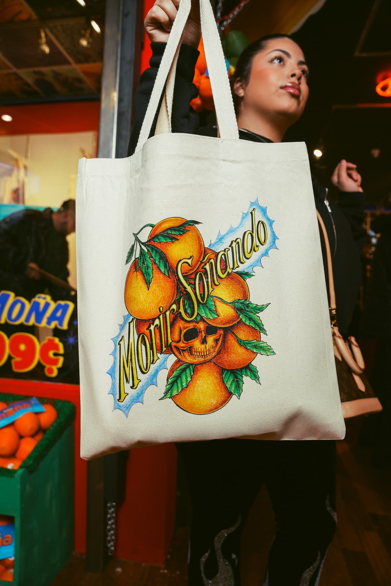 A canvas tote printed with the oranges-and-skull art held up by a guest, a 'MOÑA 99¢' window sign and an orange crate behind
