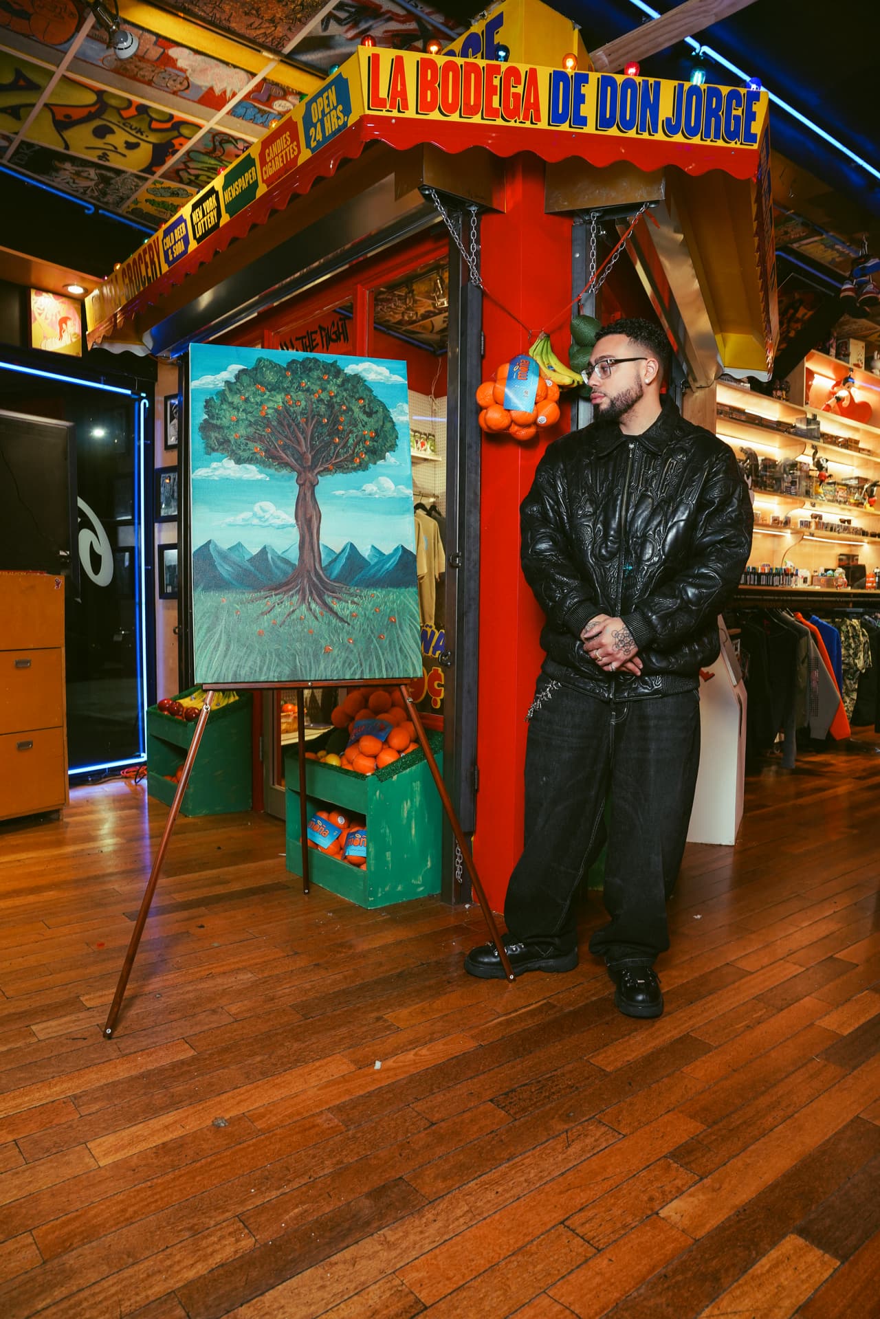 A guest beside an easel holding an original painting of an orange tree over mountains, an orange crate in the foreground