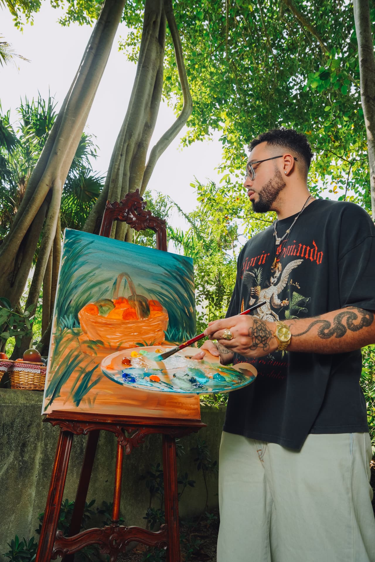 Man in the cherub tee painting an orange-basket canvas at an easel under a banyan tree, palette in hand