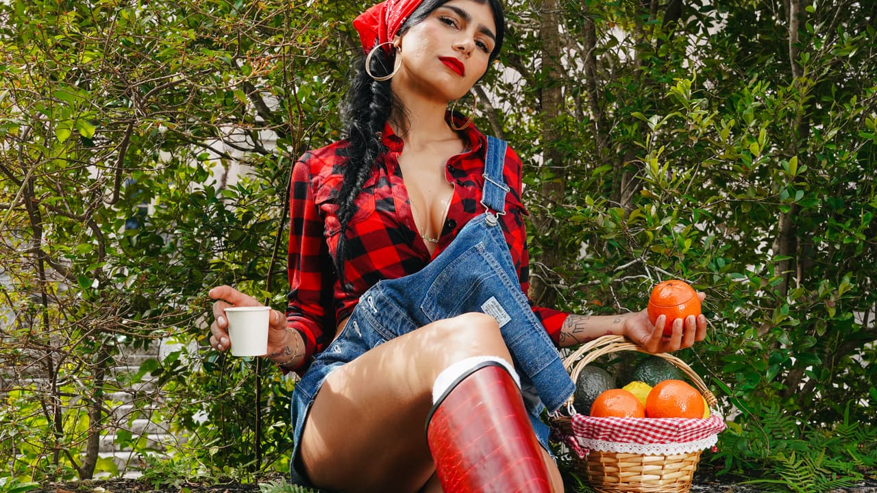 Woman in a red bandana and red boots seated on a mossy stone wall over a coral-pink wall, holding a paper cup