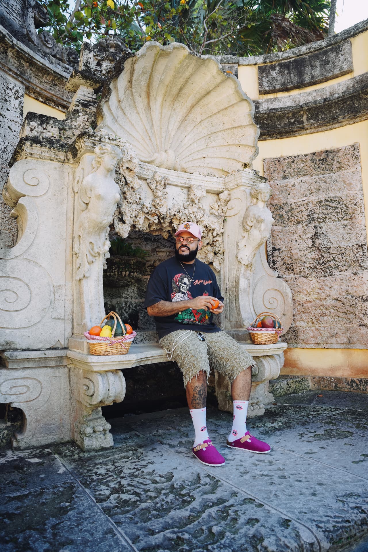 Man in a pink cap and the harvest tee seated on an ornate stone bench in a shell grotto, fruit baskets on either side