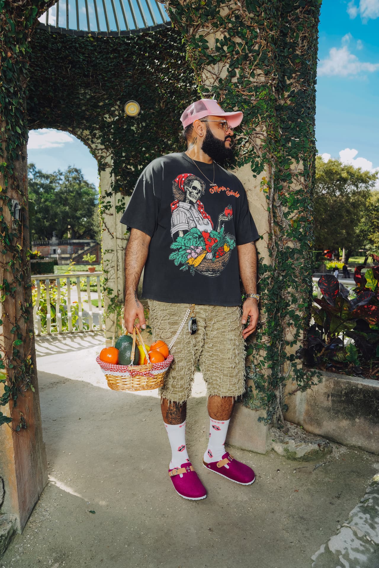 Man in a pink trucker cap and the harvest tee standing under an ivy-covered stone arch with a fruit basket