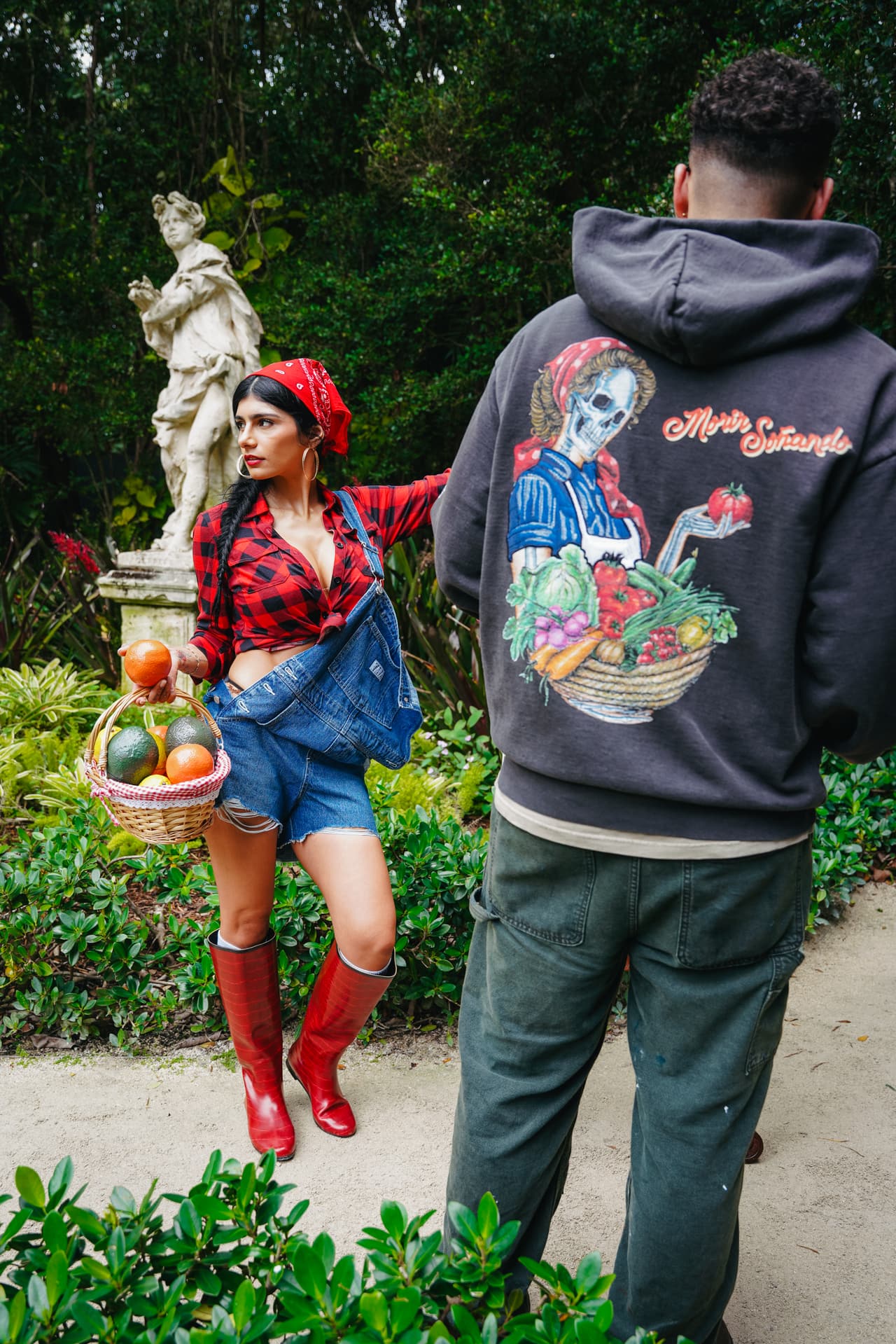 Woman holding an orange toward a garden statue while a man in the harvest hoodie stands with his back to camera, showing the hoodie's back art