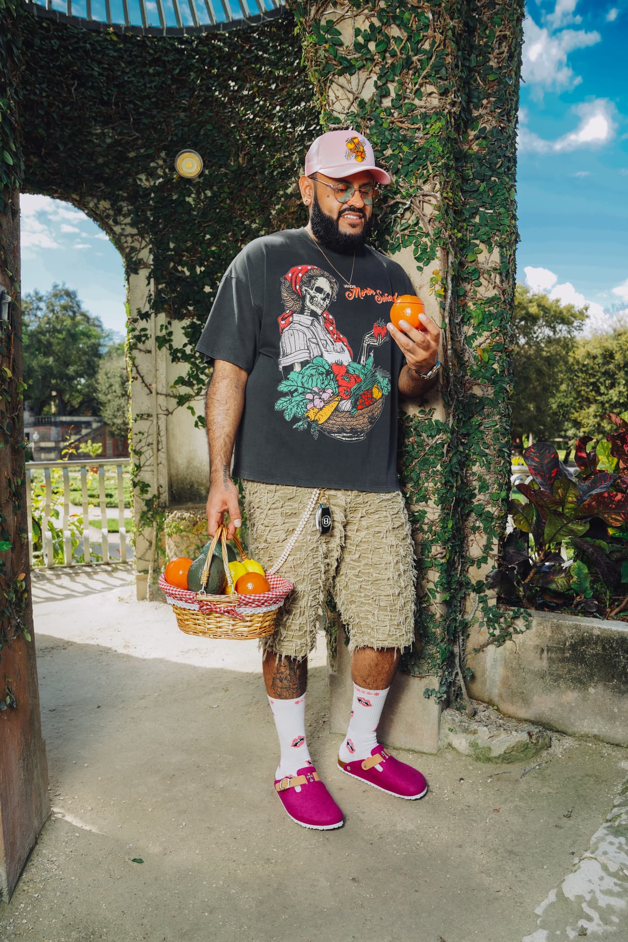 Man in a pink trucker cap and the harvest tee smiling while holding an orange-shaped jar up to the camera under a stone arch