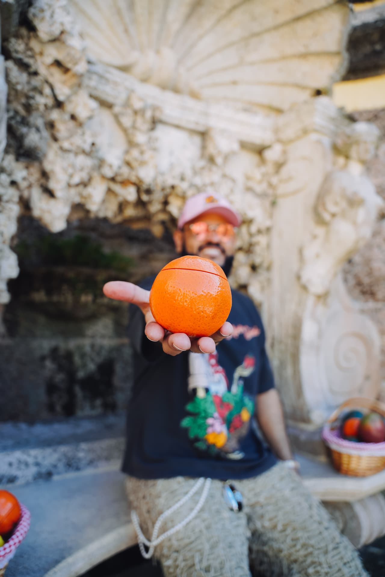 Close detail of a hand holding an orange-shaped jar to camera, a man in a pink cap out of focus behind in a shell grotto