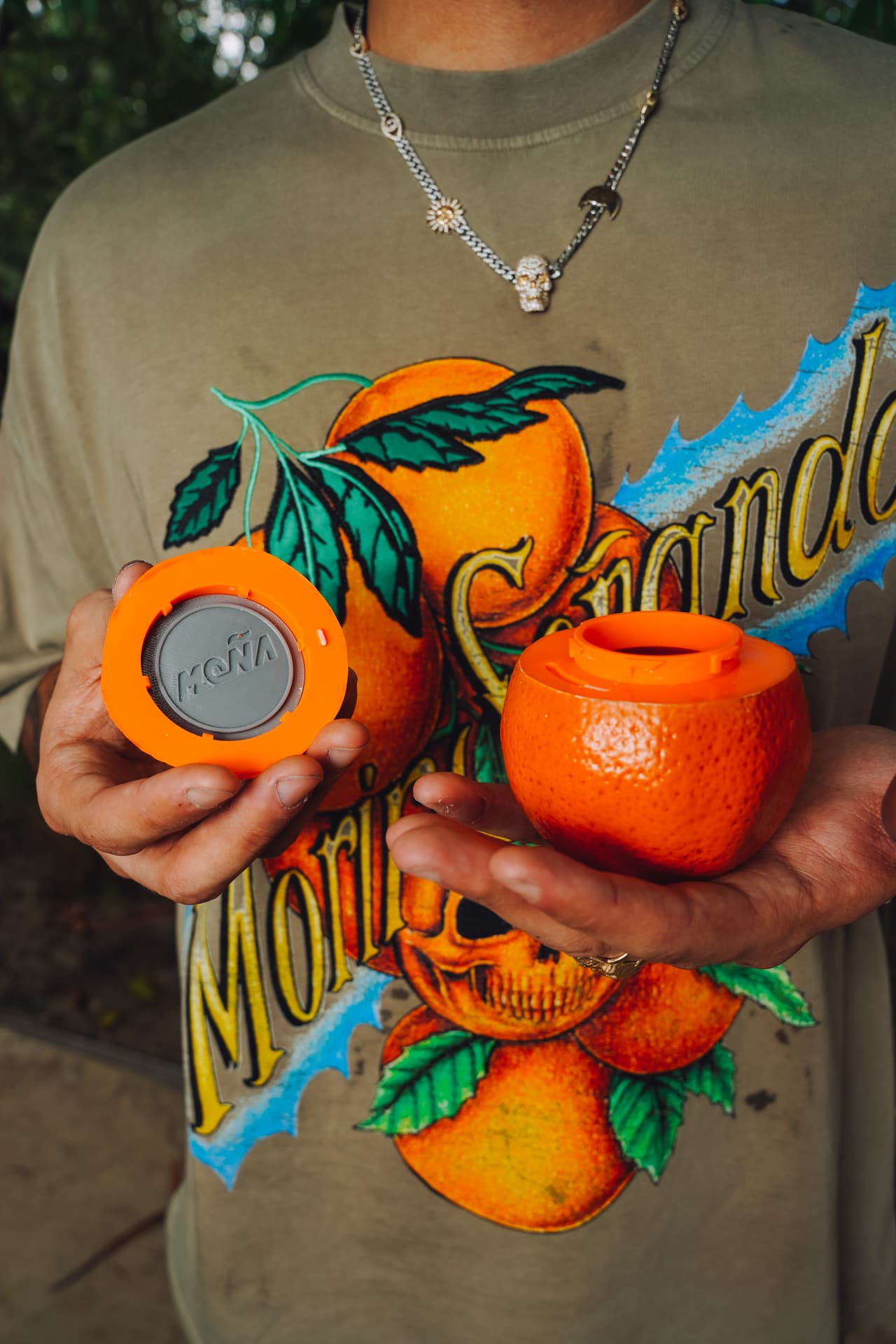 Hands holding an open orange-shaped jar with a grey embossed MOÑA lid, over a tan oranges-and-skull tee