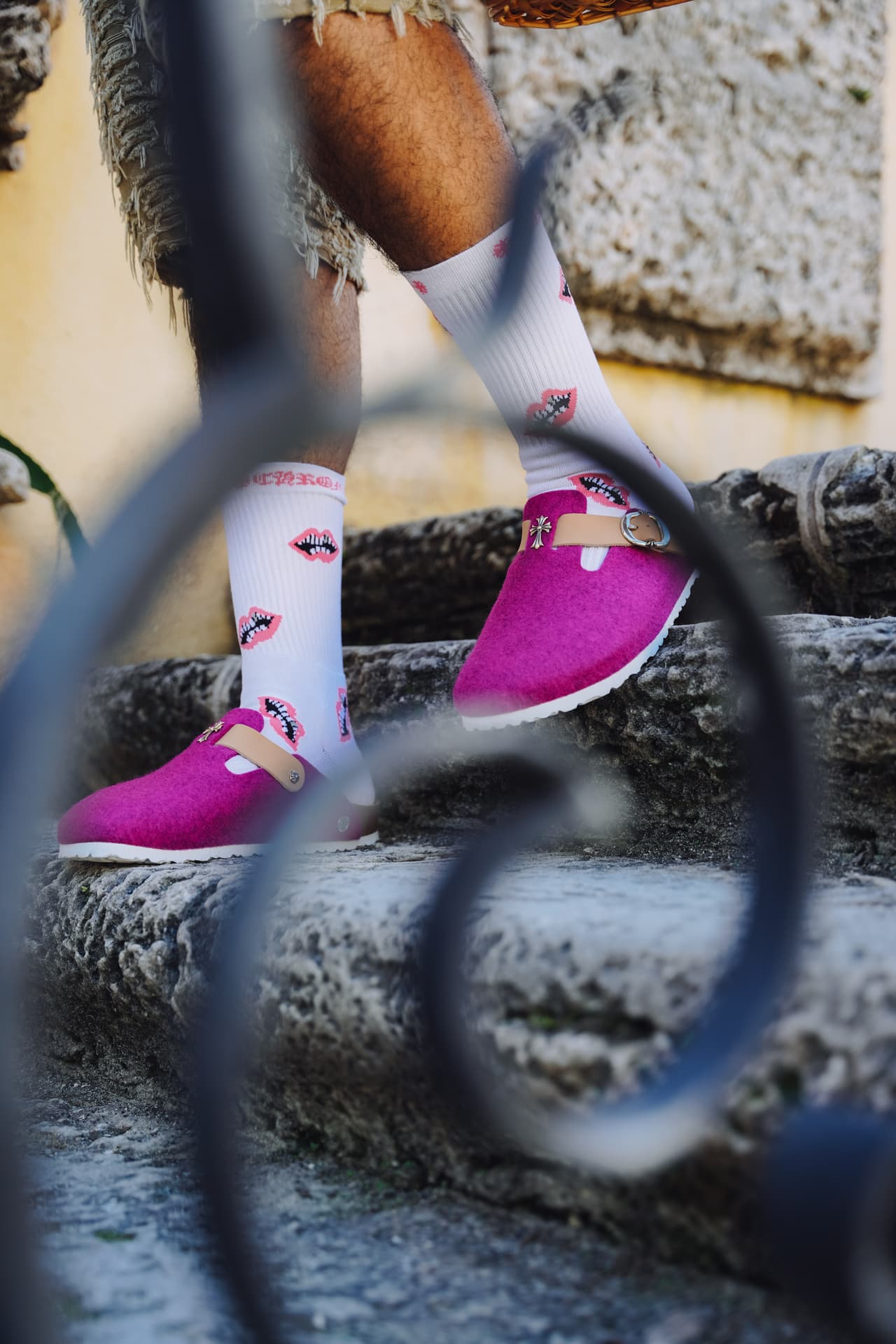 Detail through an iron scroll railing of fuchsia clogs and lips-print socks on stone steps