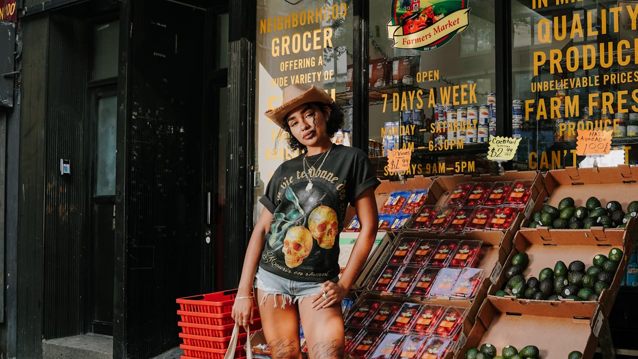 Model in a straw cowboy hat and the lemon-skulls tee standing centered under the full STILES FARMERS MARKET awning, a tote at her side