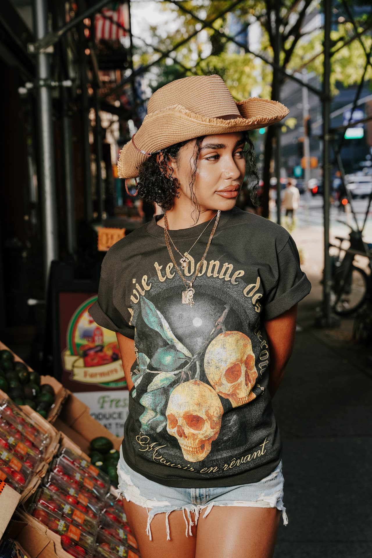 Close portrait of the model in the lemon-skulls tee, gold chains, looking to the side against a sidewalk scaffold