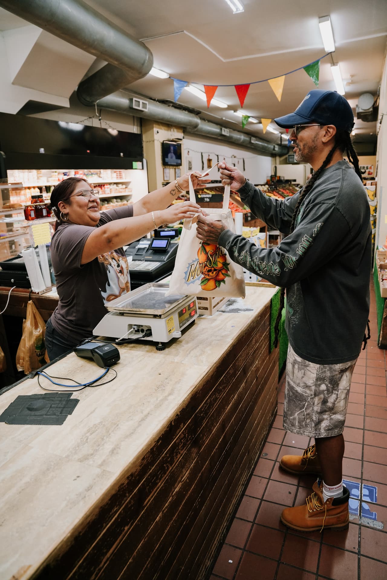 A bodega cashier handing an oranges-print tote across the counter to a man in a navy cap