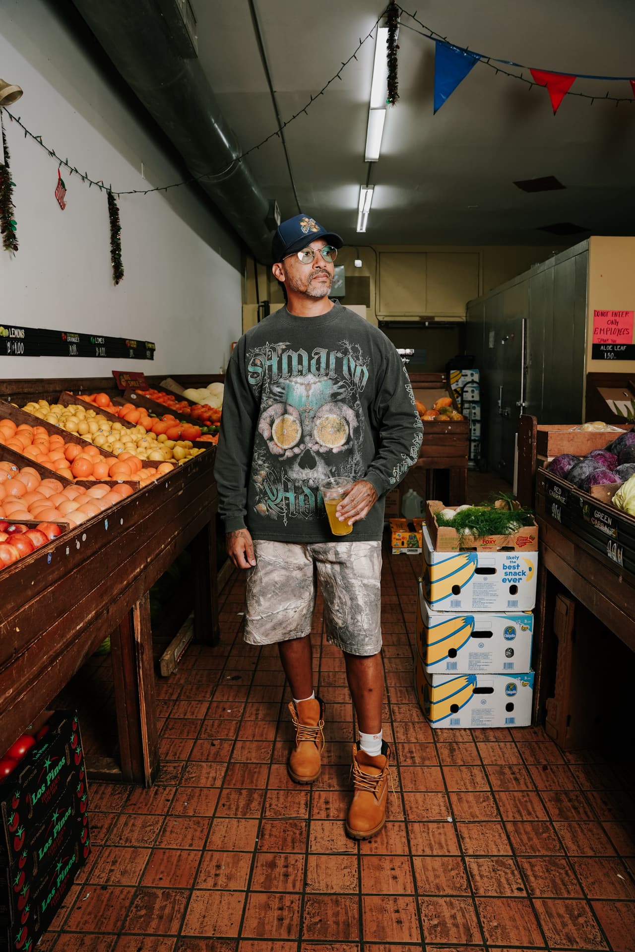 Man in a navy cap, long-sleeve tee and camo shorts standing in a bodega produce aisle with a juice cup