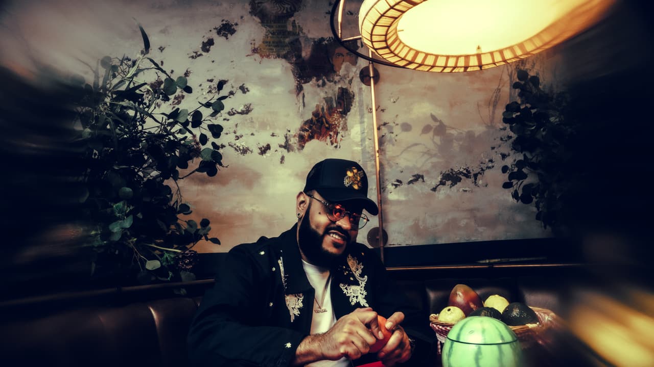 Man in a black cap and embroidered black jacket at a red-clothed table with watermelon, mangos and lemons; a painted-foliage wall, hanging lamp and teal-red bar light behind