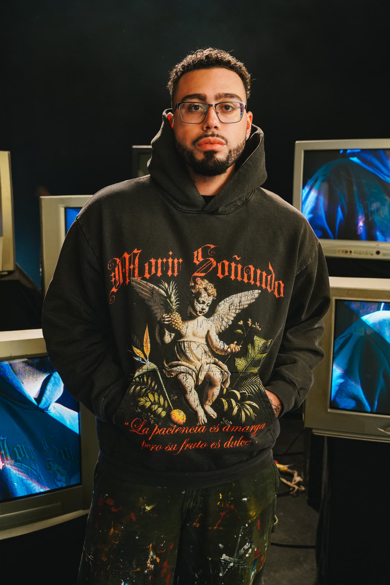 Waist-up in front of a wall of switched-on television sets, the cherub hoodie's blackletter lockup and winged cherub reading whole