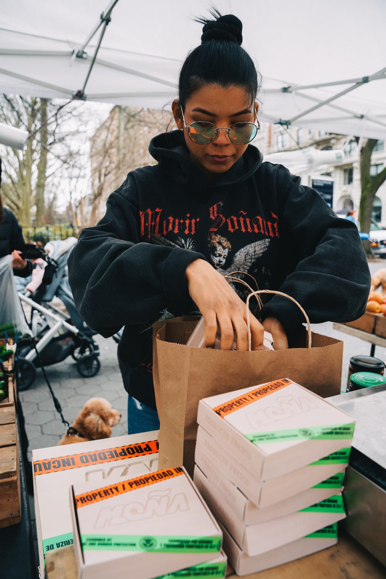 Woman in the cherub hoodie packing stencilled Moña crates into a paper bag at a winter market stall