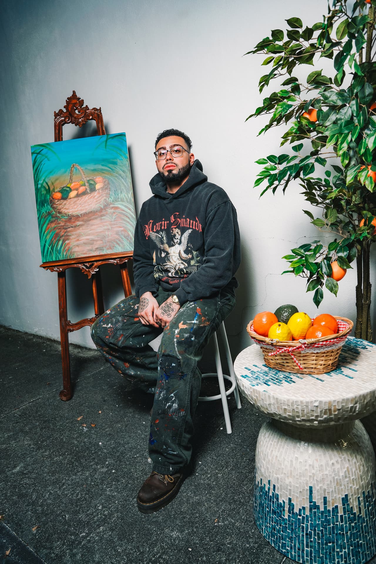 Man in the cherub hoodie seated on a stool between a finished fruit-basket painting and a potted orange tree