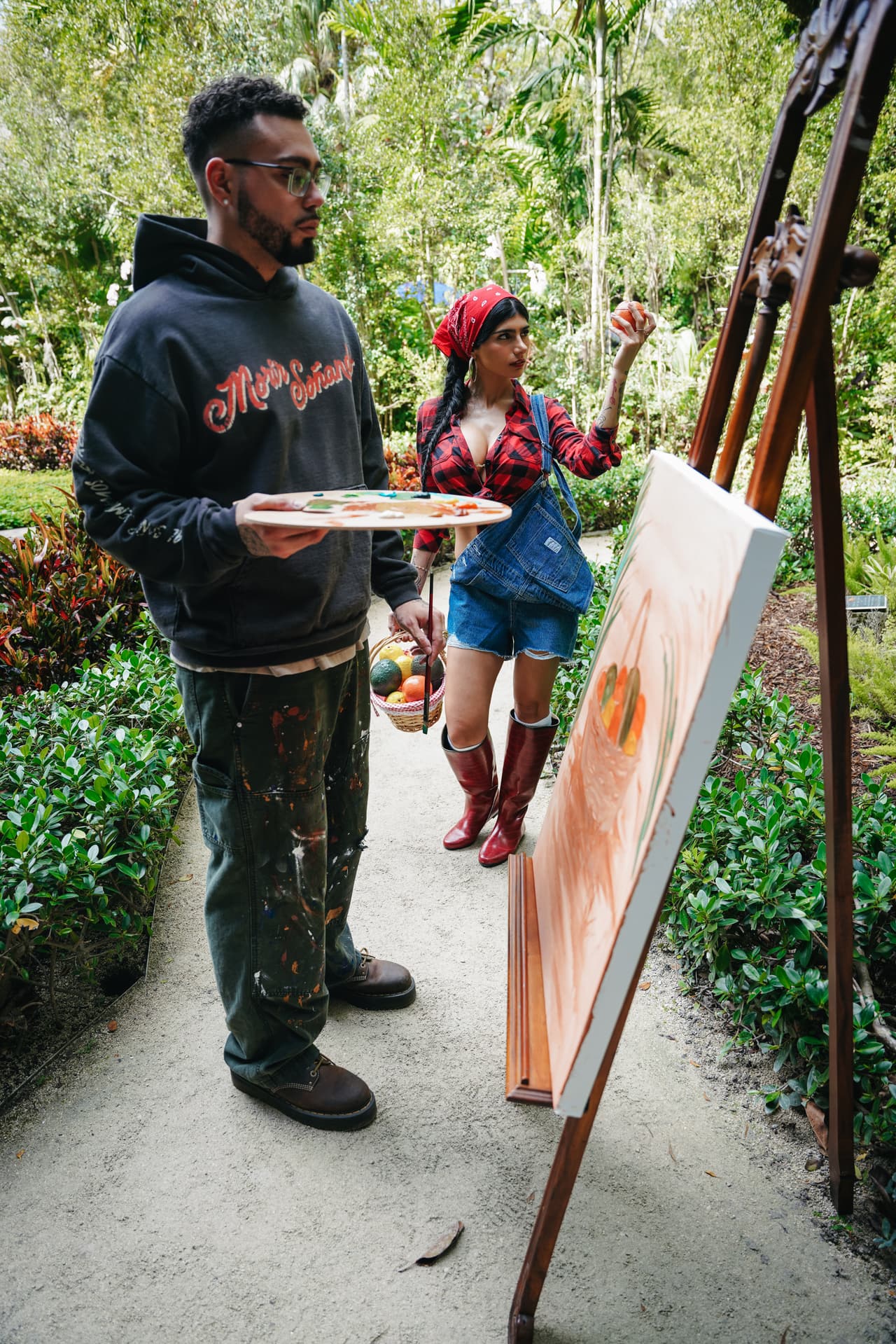 Man standing at a garden easel in the harvest hoodie with a loaded palette, the red script across his chest