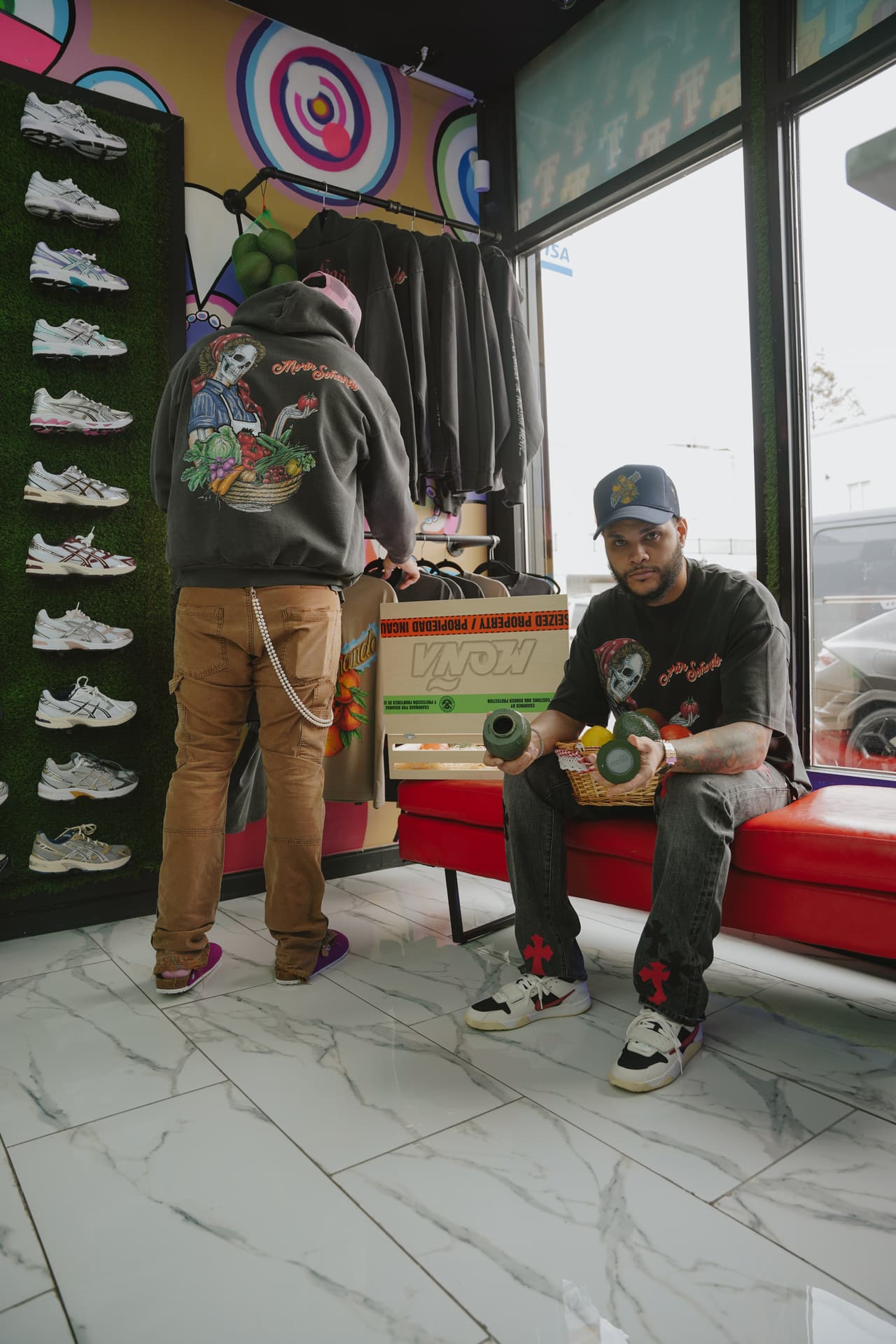Shopper facing a merch rack in a sneaker store, the harvest hoodie's back print whole across his shoulders and a pink trucker cap under the hood