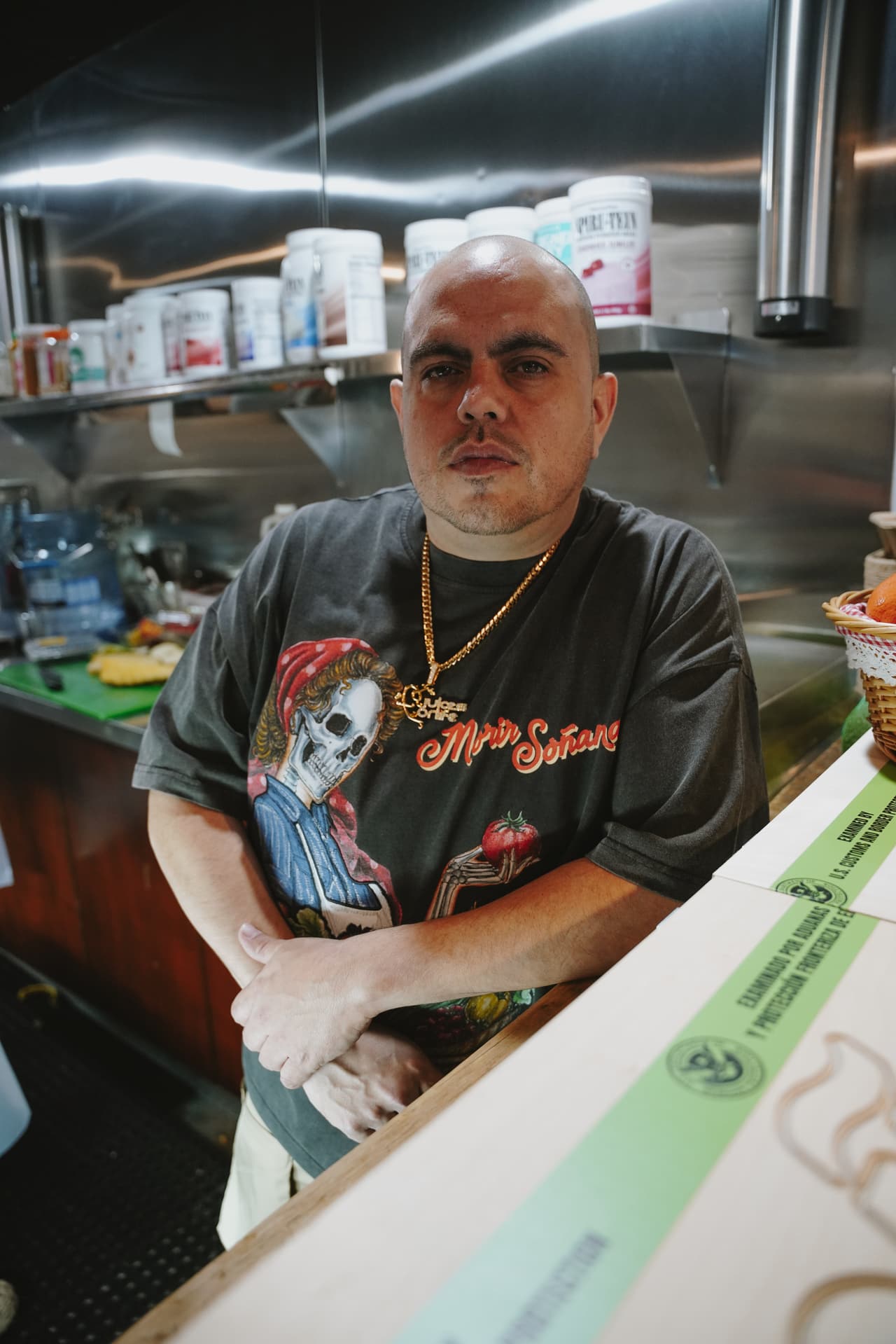 Man behind a juice-bar counter in the harvest tee, the skeleton harvest woman square to camera