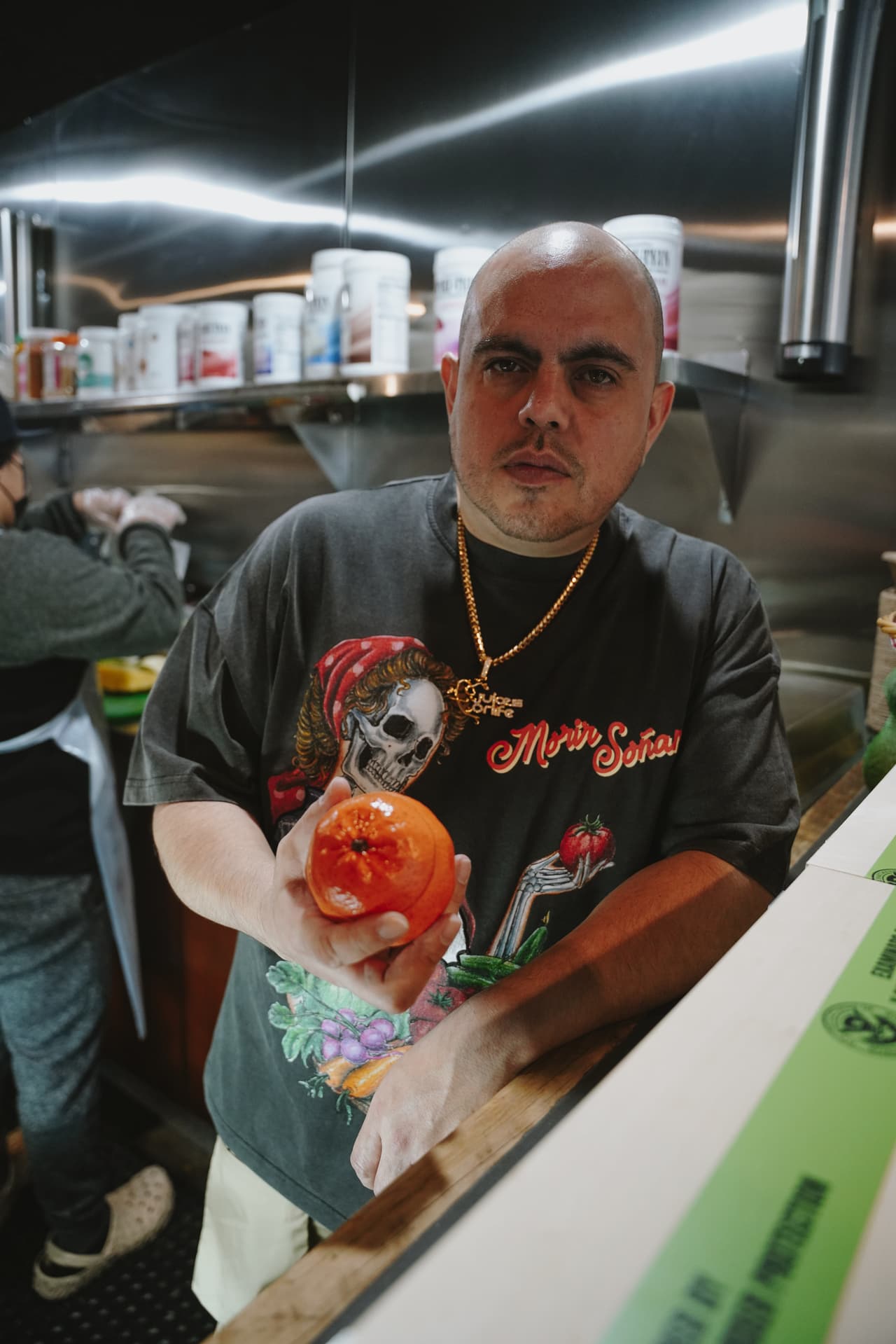 Man in the harvest tee holding a ceramic orange out across a steel juice-bar kitchen