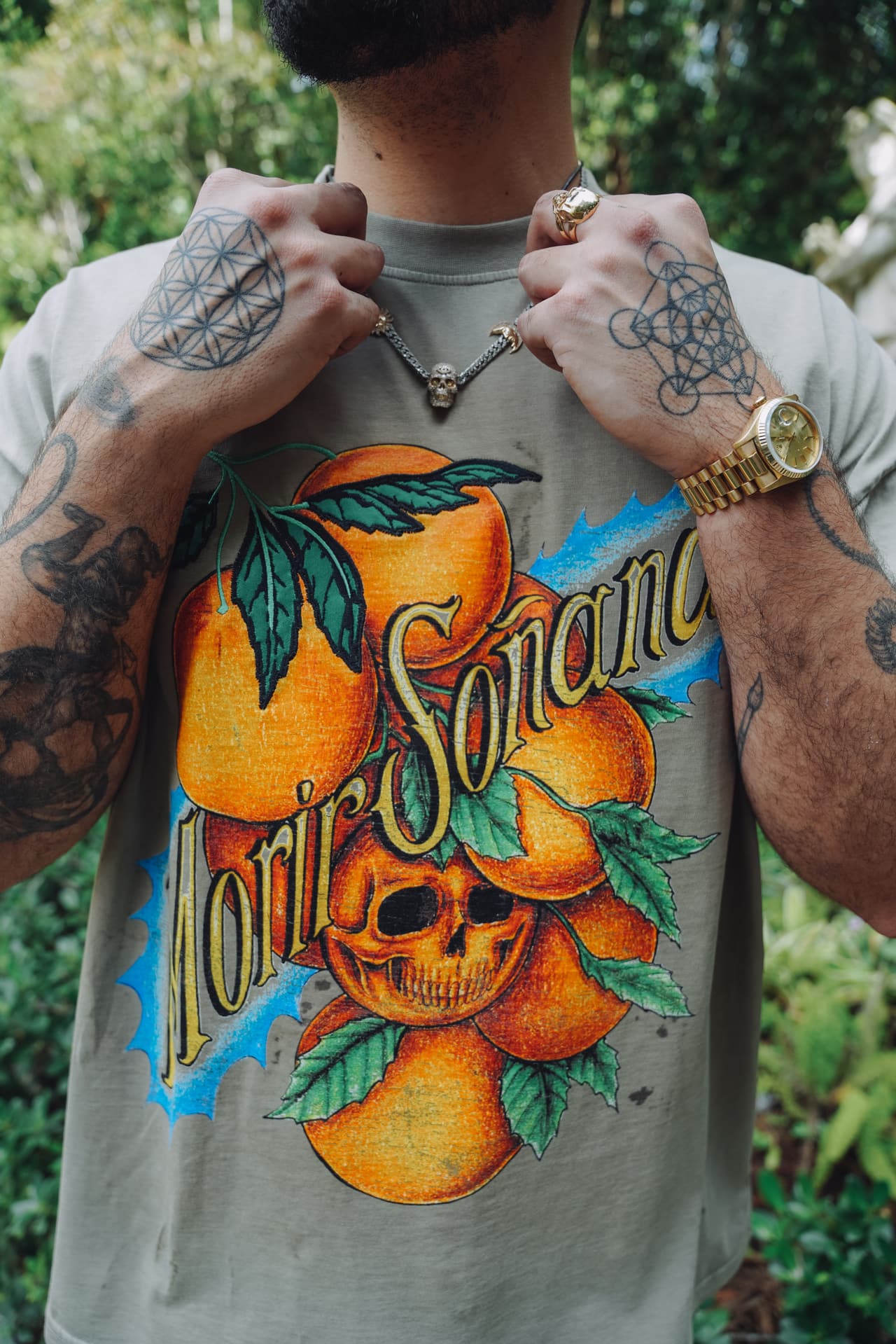 Chest crop of the brown tee with both hands lifting a chain clear, the oranges, leaves and skull filling the frame