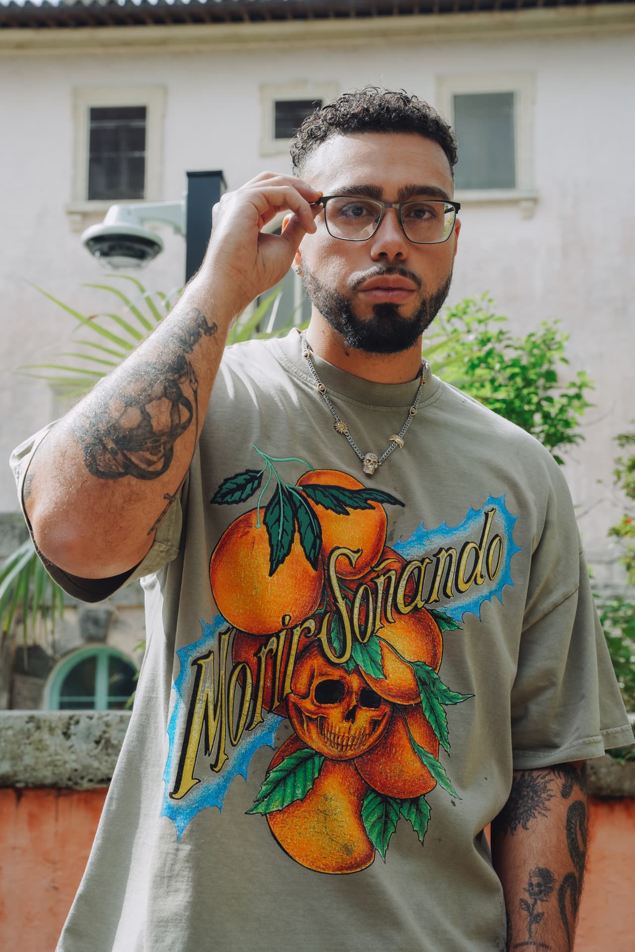 Man lifting his glasses in front of a villa wall, the oranges-and-skull print at catalogue scale across the brown tee