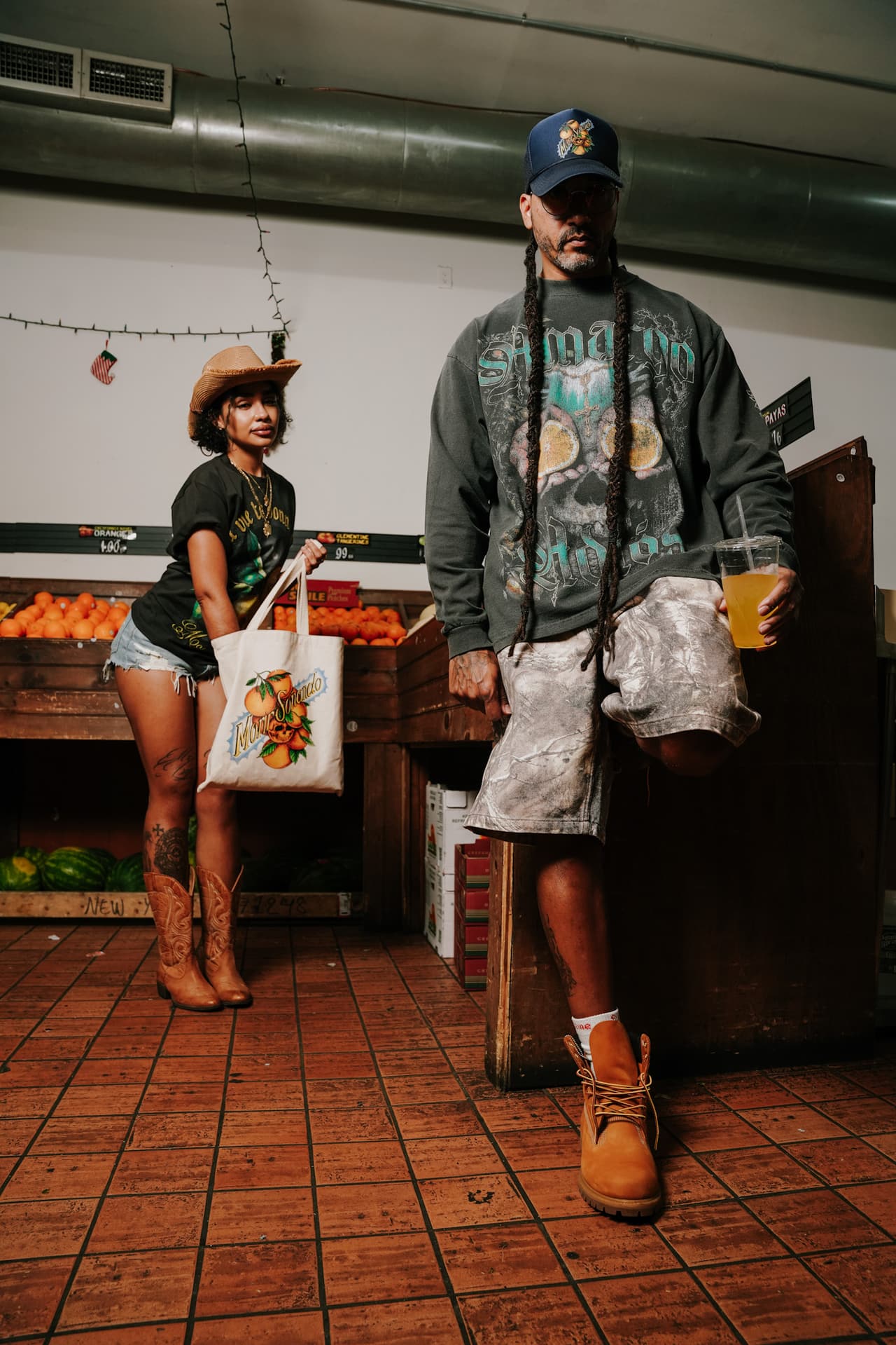 Two shoppers inside a produce market, the canvas tote hanging from her hand beside a crate of oranges